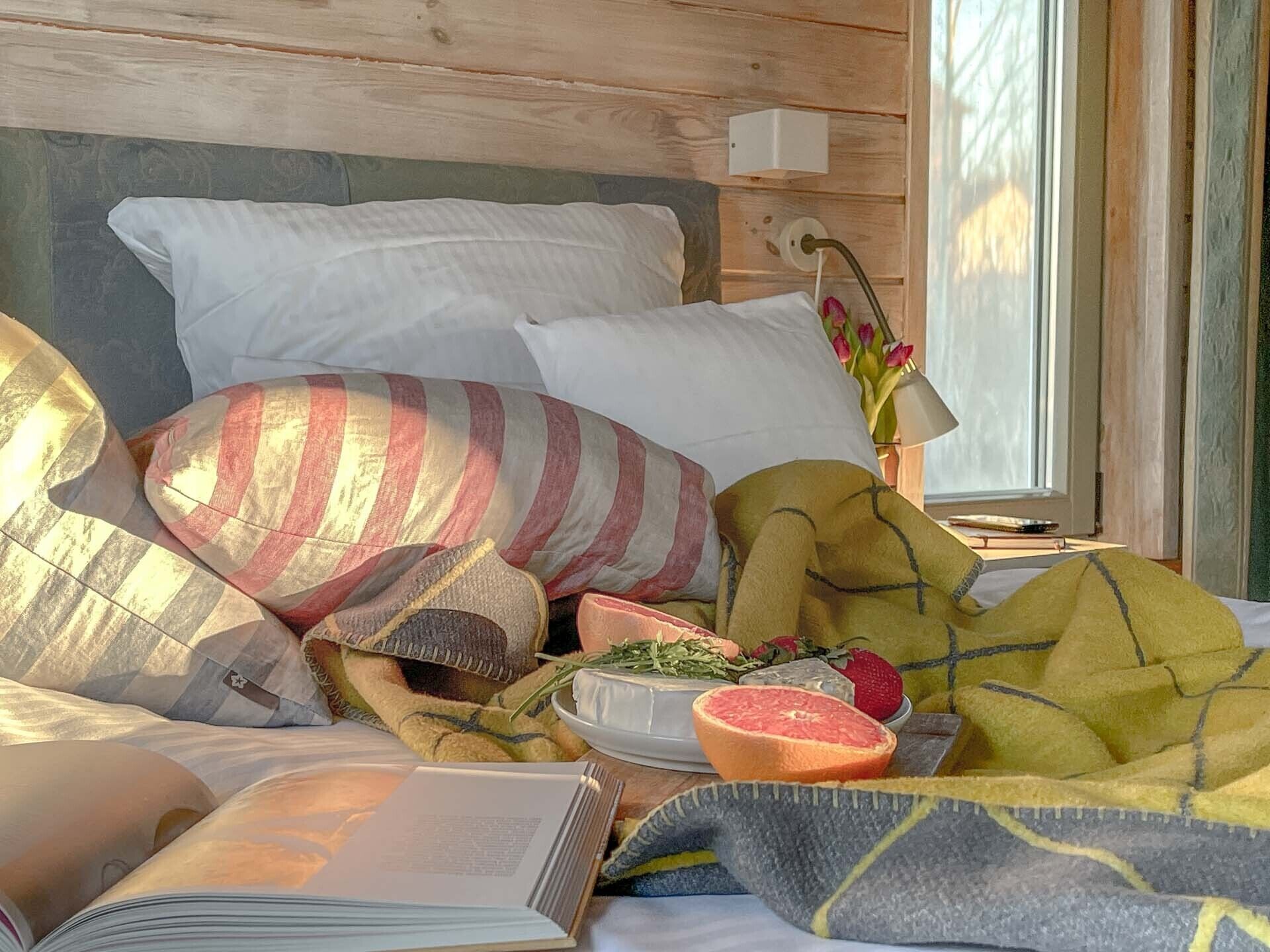 A cozy bedroom with a bed, pillows, a bedside table with a lamp, a window with a view of trees, and a book on the bed.