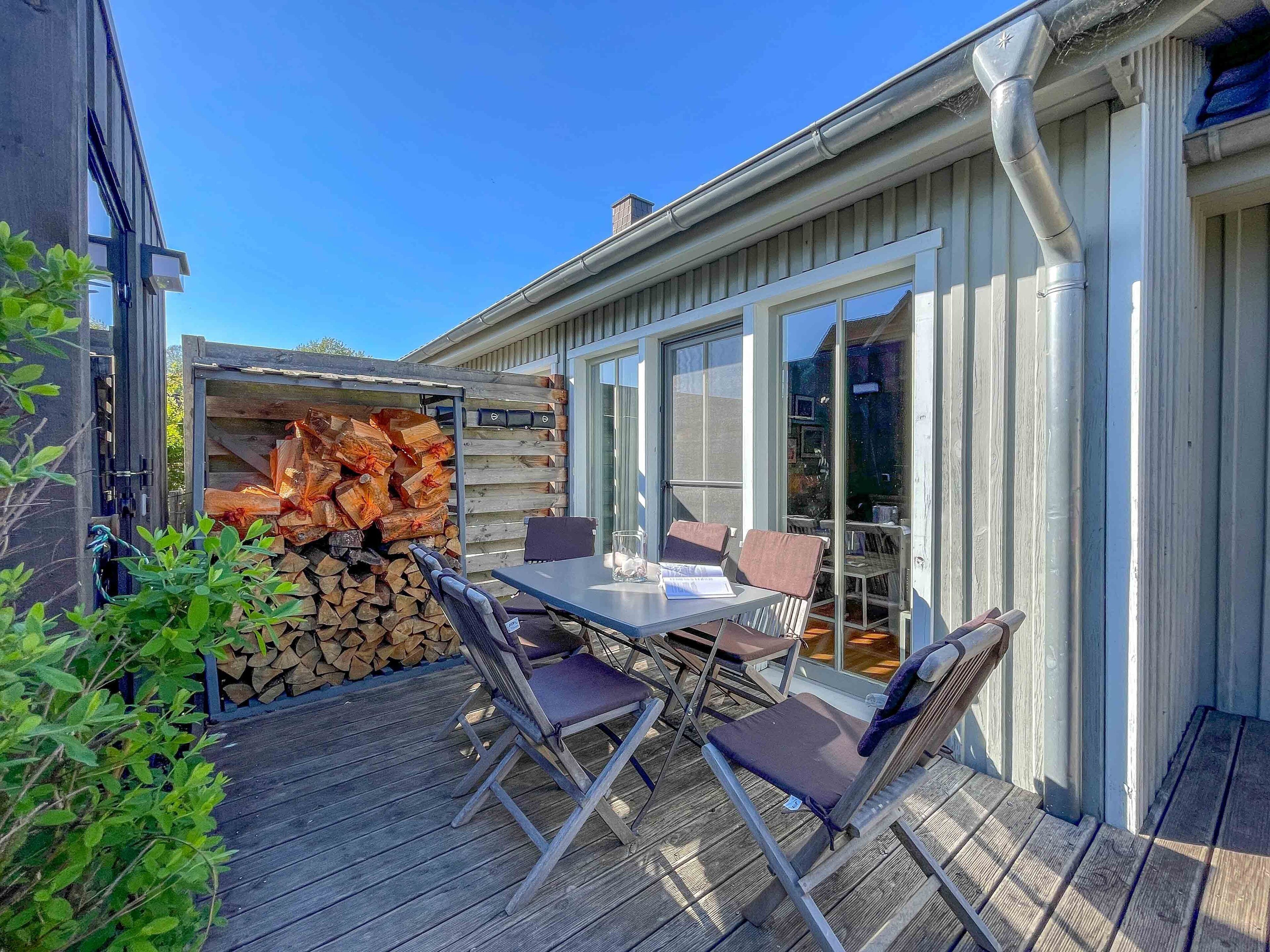Een terras buiten met een tafel, stoelen, een stapel houtblokken en een glazen deur die naar een kamer leidt.