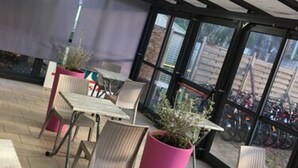 Un patio interior con mesas y sillas, plantas grandes en macetas y vistas a motocicletas estacionadas afuera.