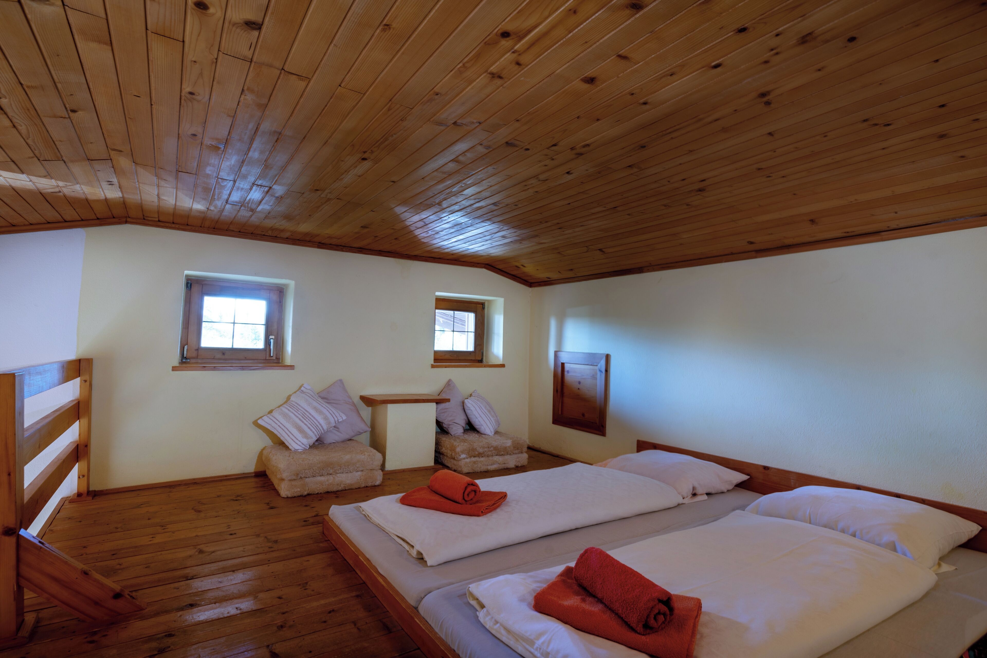 A room with wooden floors and ceiling, two beds with white linens and orange towels, two chairs with cushions, and two windows.