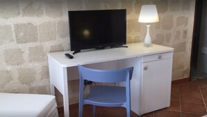 Ein kleiner Raum mit einem weißen Schreibtisch, einem schwarzen Fernseher, einem blauen Stuhl und einer weißen Lampe.