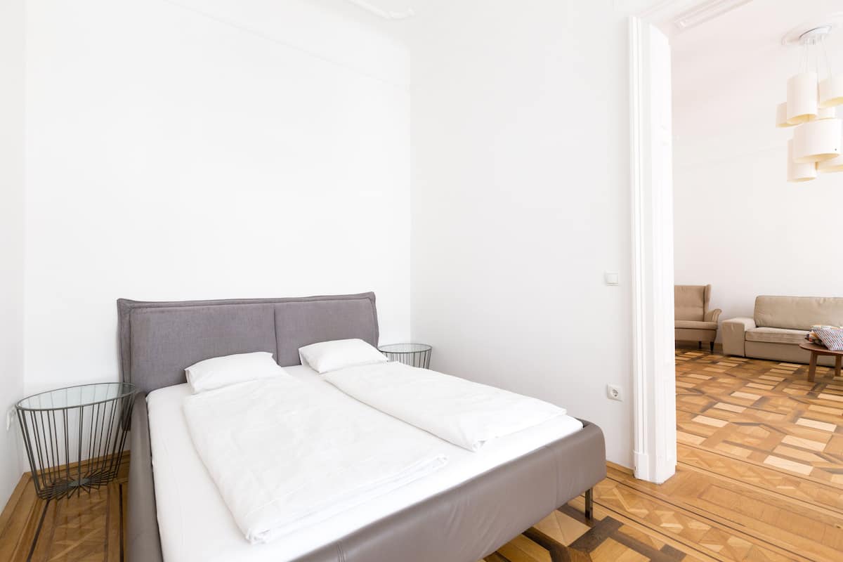 A neatly made bed with white linens, a grey headboard, and a small bedside table with a lamp.