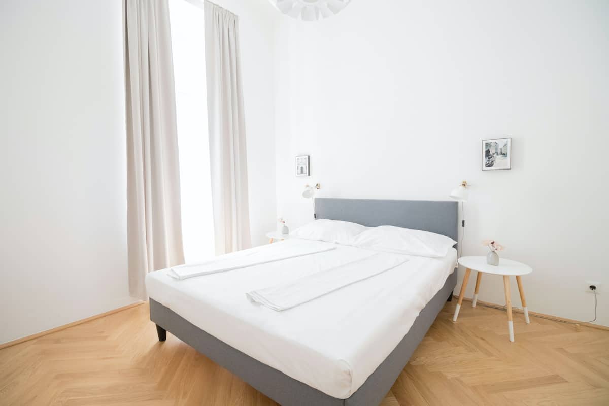 A modern bedroom with a grey bed, white linens, a small bedside table with a lamp, and a picture on the wall.