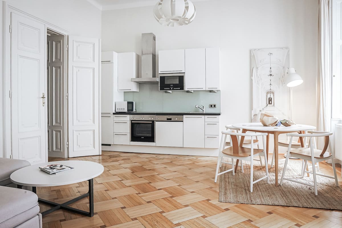 A modern kitchen with white cabinets, a built-in oven, and a microwave. There is a dining area with a round table and chairs.