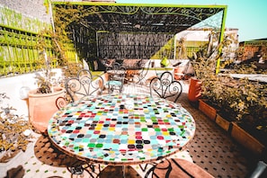 Una mesa redonda con un tablero de mosaico colorido, rodeada de sillas de metal, en un patio al aire libre con plantas en macetas y una valla metálica verde.
