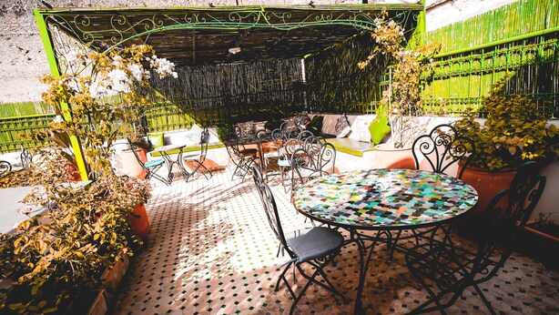 Un patio al aire libre con una mesa redonda y sillas, rodeado de plantas en macetas y una valla metálica verde.