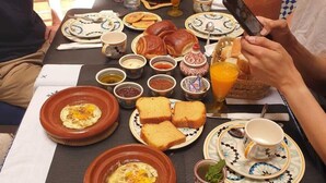 Una mesa de comedor con diferentes alimentos para el desayuno, como pan, huevos y jugo de naranja, ubicada en una habitación con baldosas estampadas y una gran maceta de barro.