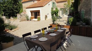 Un área de comedor al aire libre con una mesa preparada para cuatro personas, rodeada de sillas, en un patio con una casa al fondo.