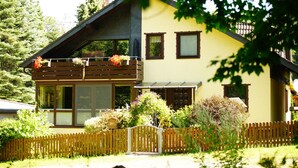A two-story house with a balcony, a wooden fence, and various plants. - Ferienwohnung im Wald mit Kamin (Bad Harzburg)