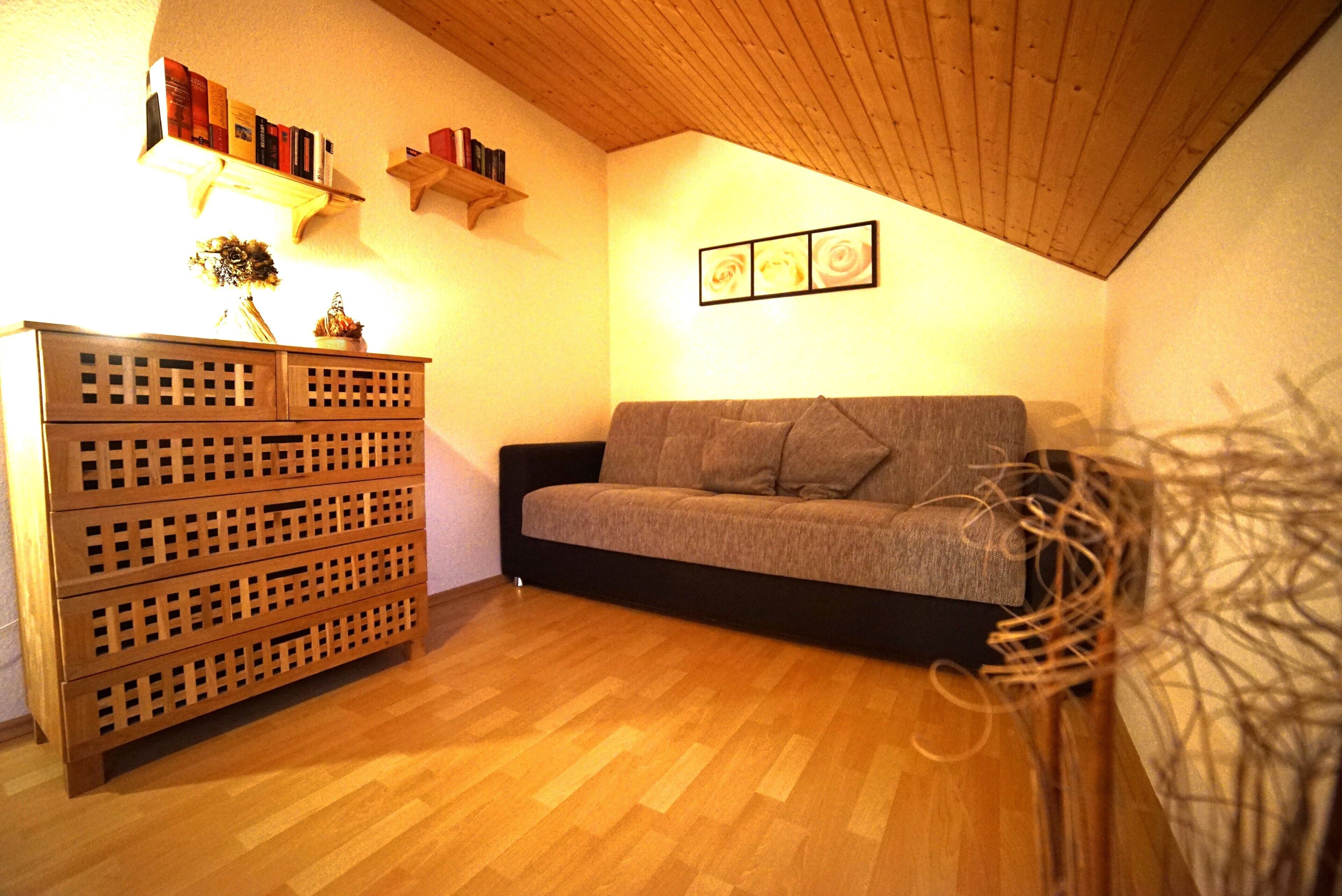 A cozy living room with a wooden floor, a grey sofa, a wooden cabinet, and a ceiling with wooden beams.