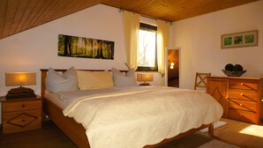 A bedroom with a large bed, two bedside tables with lamps, a wooden dresser, a window with curtains, and a painting on the wall. - Ferienwohnung im Wald mit Kamin (Bad Harzburg)