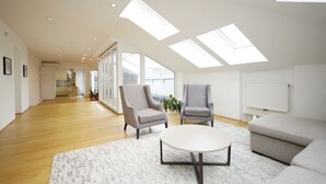 Amplia sala de estar con techo alto, tragaluces, una mesa de centro redonda, dos sillones, un sofá y vistas a una cocina y comedor a través de grandes ventanales.