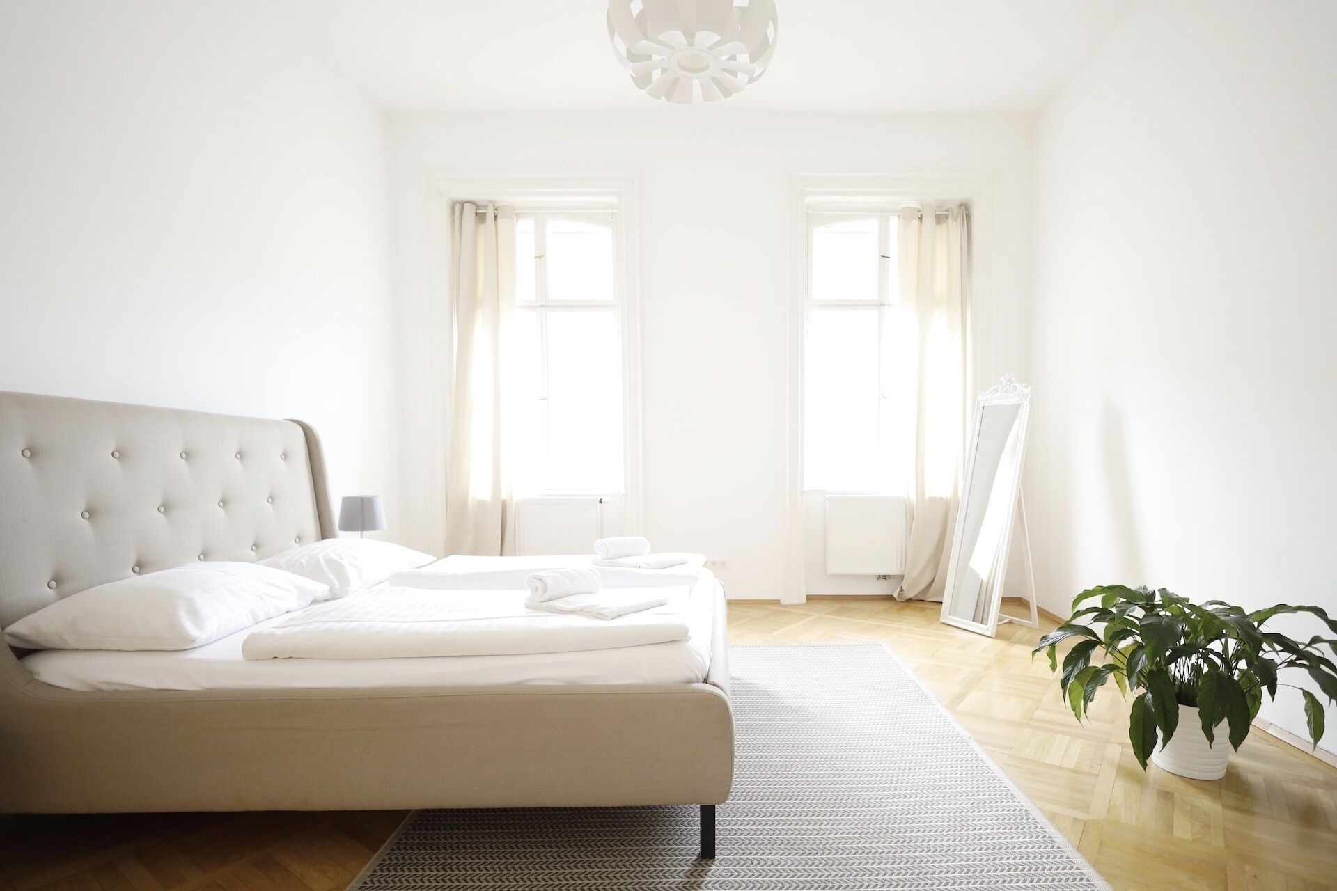 Een slaapkamer met een bed, een nachtkastje, een spiegel, een plant en twee ramen.