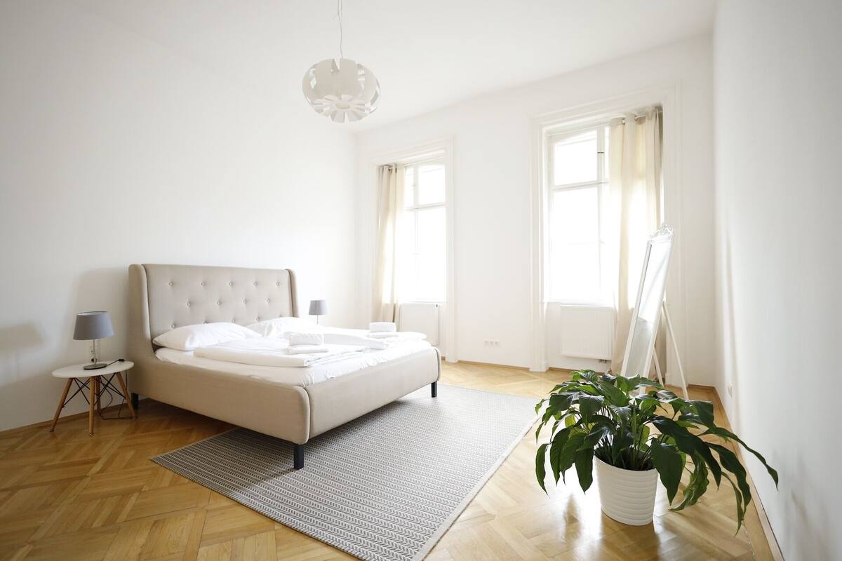 A minimalist bedroom with a bed, bedside lamps, a hanging light fixture, a radiator, and a potted plant.