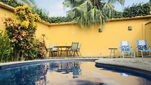 Una piscina con borde de azulejos, rodeada por un muro amarillo, y mobiliario exterior con sillas y una mesa.