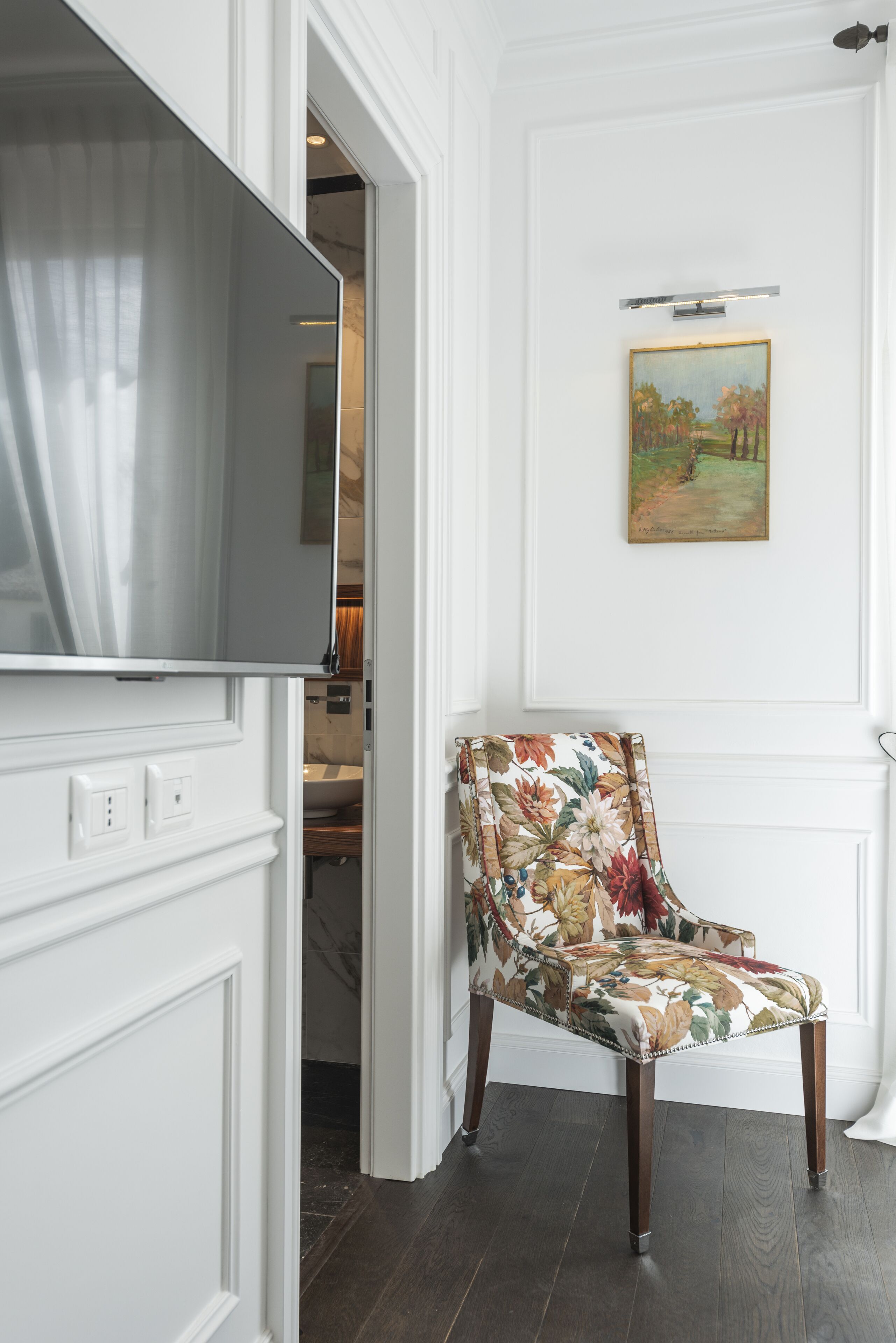 Una habitación con televisión de pantalla plana montada en una pared blanca, un sillón estampado con motivos florales y un cuadro sobre una puerta.