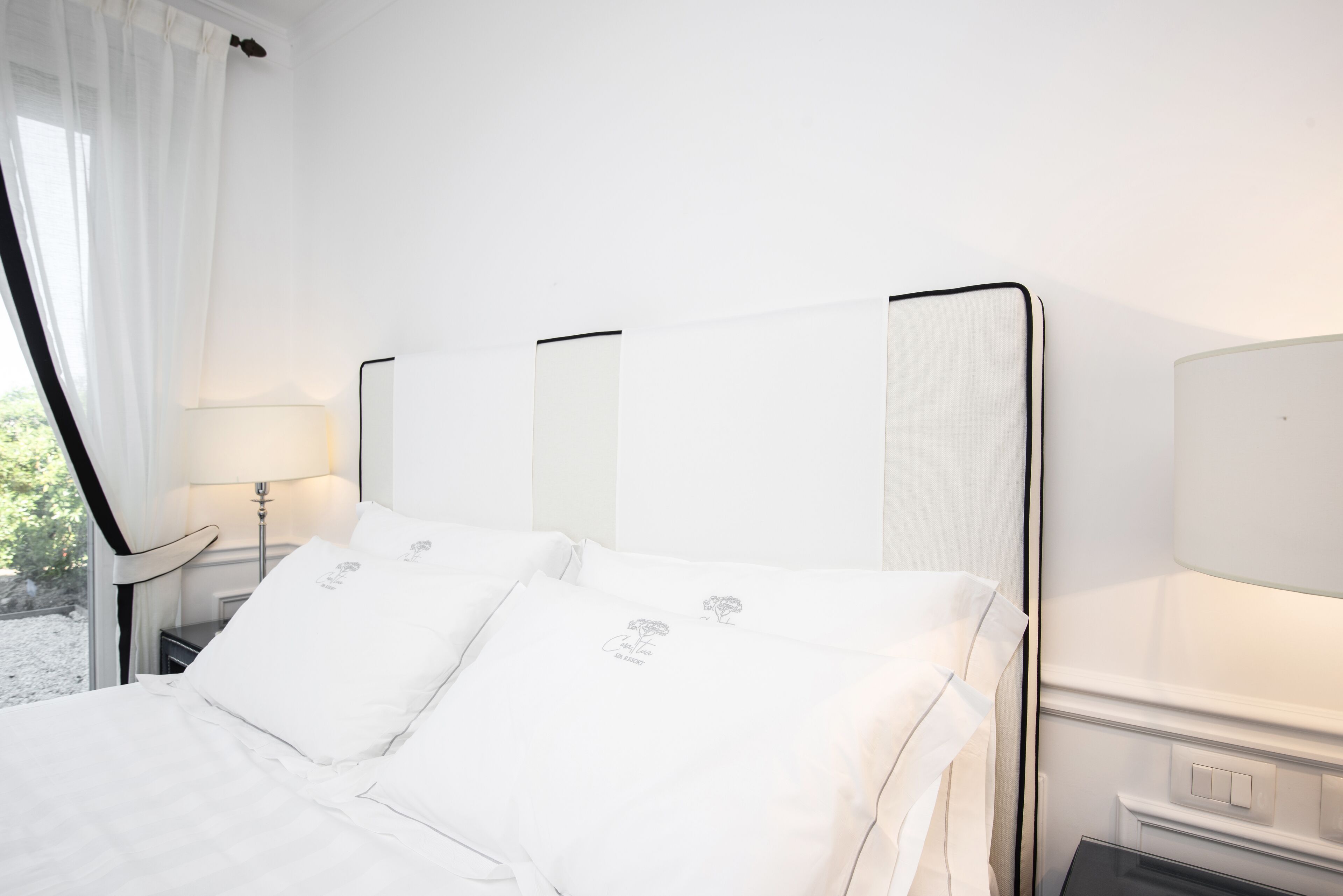 Una cama bien tendida con sábanas y almohadas blancas, una mesita de noche con una lámpara, y una ventana con cortinas traslúcidas.