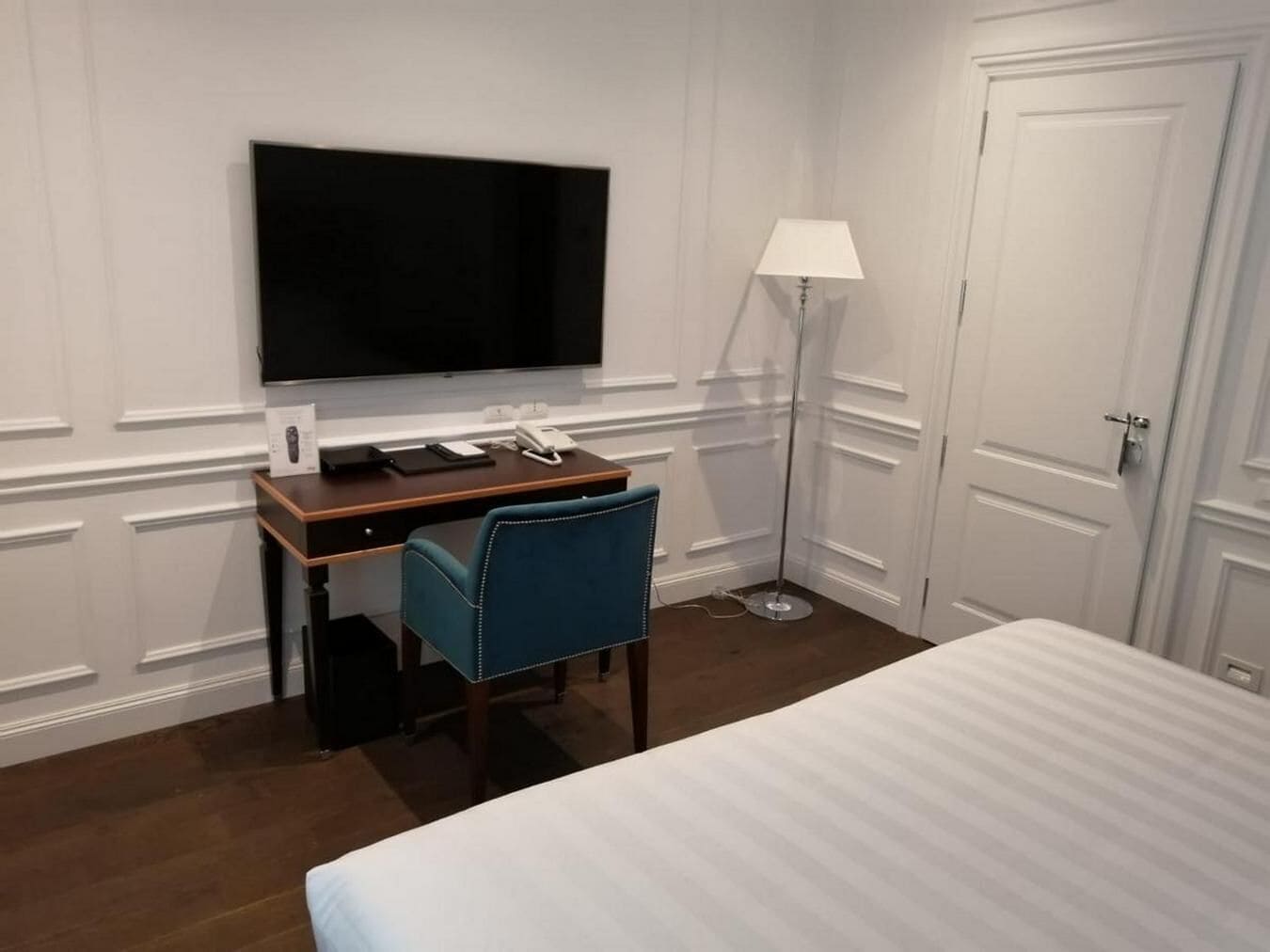 Een hotelkamer met een bed, een bureau met een flatscreen televisie, een stoel, een lamp en een deur.