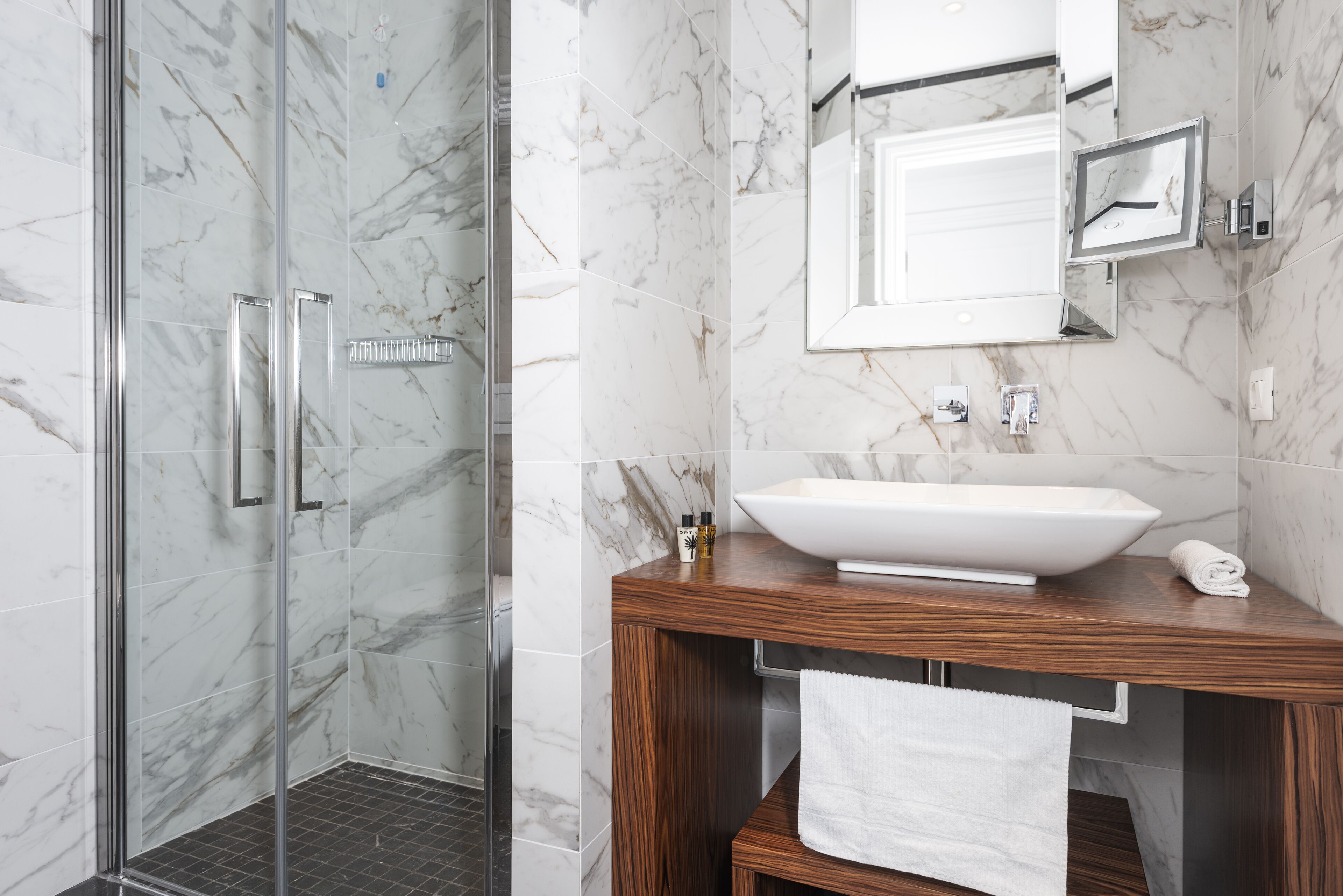 A modern bathroom with a glass shower enclosure, a wooden vanity with a white sink, and marble walls.