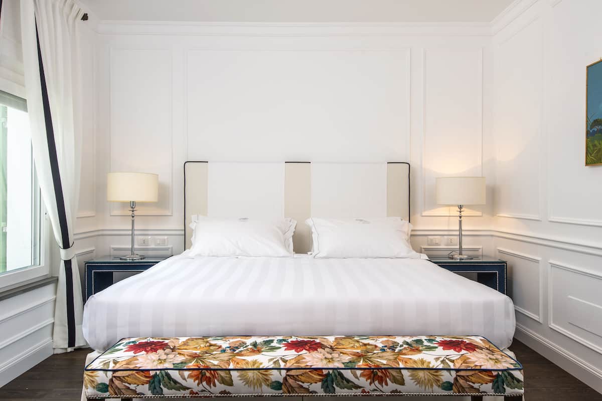 A neatly made bed with white linens, two bedside tables with lamps, and a floral-patterned bench.