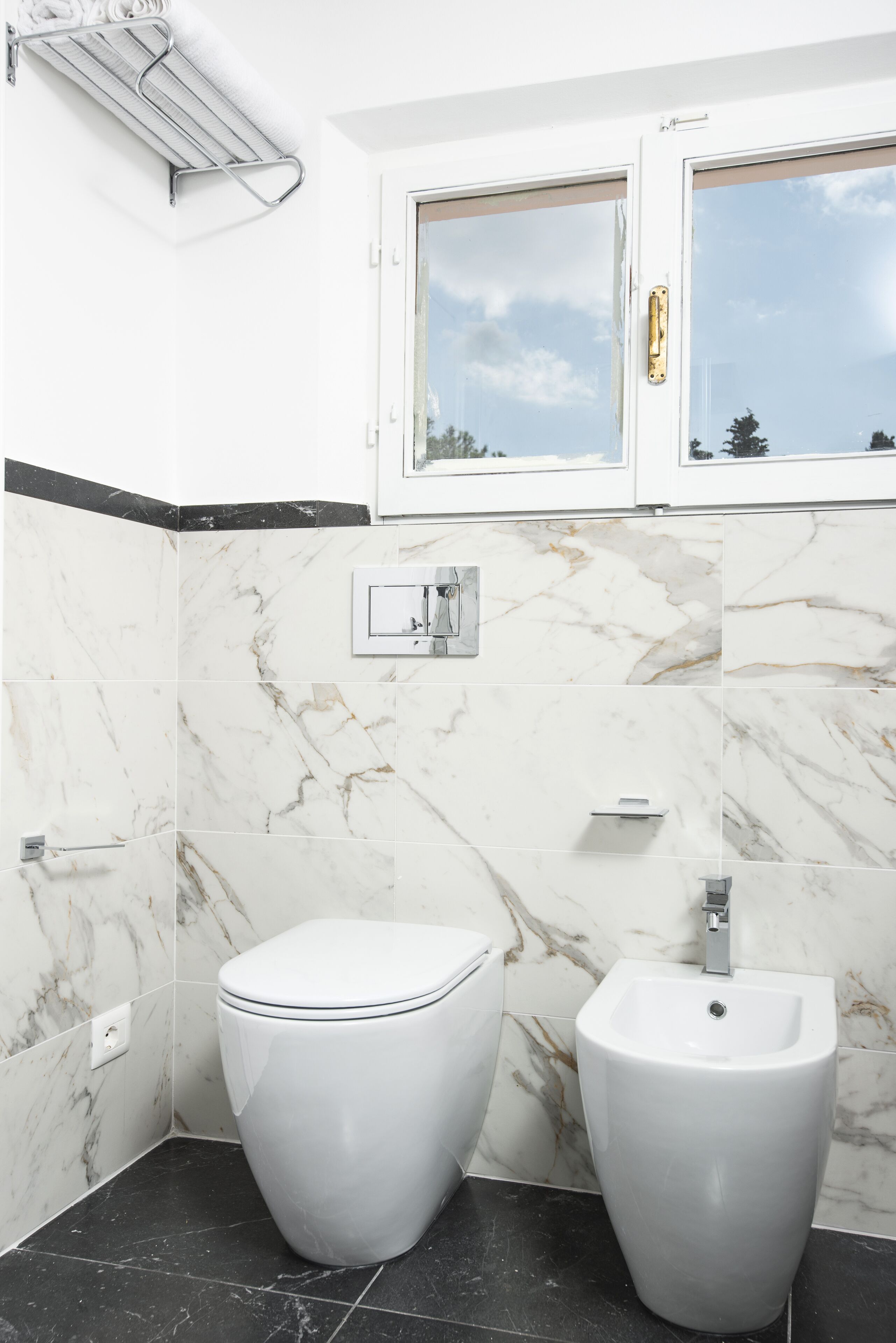 A bathroom with a toilet, bidet, and a window with a view of the sky.