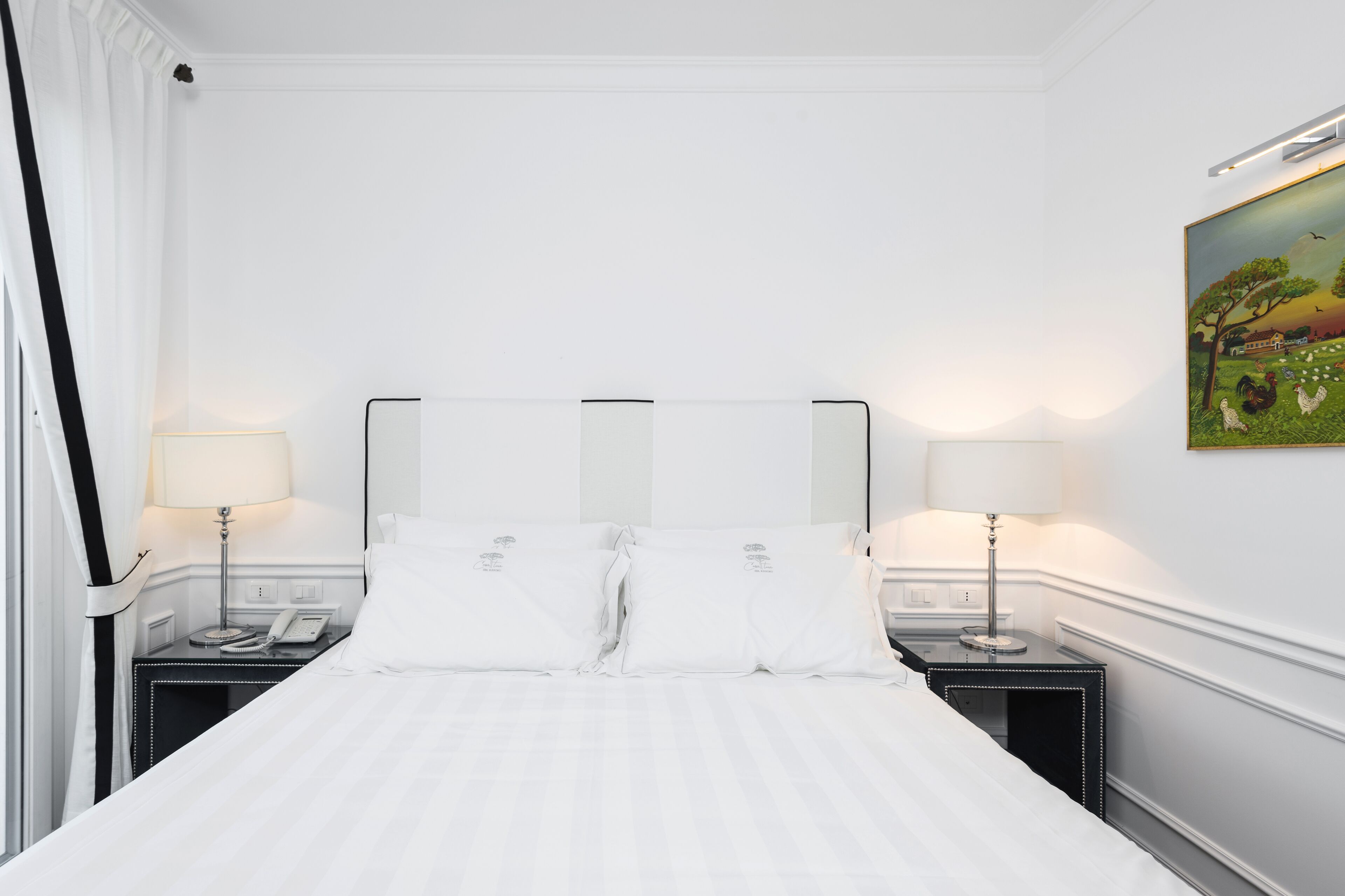 Habitación de hotel con una cama grande, dos mesitas de noche con lámparas, un cuadro en la pared y una ventana con cortinas.