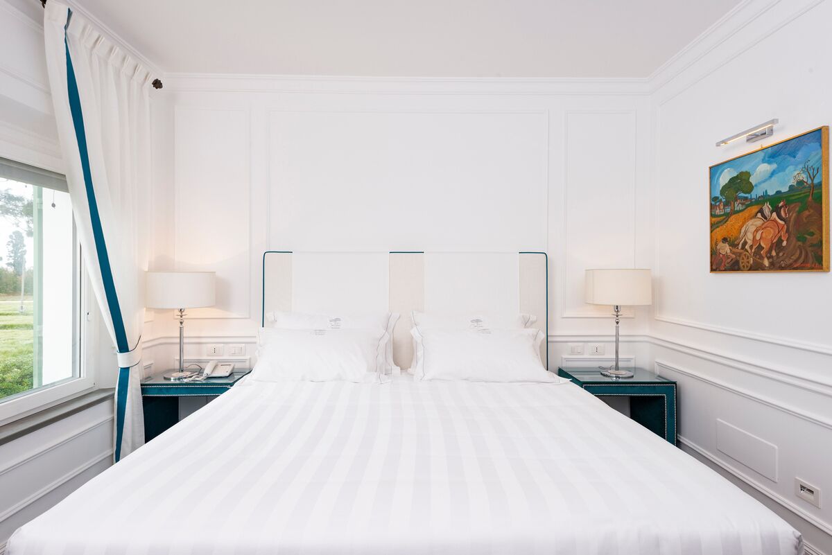 A hotel room with a large bed, two bedside tables, a window with curtains, and a painting on the wall.