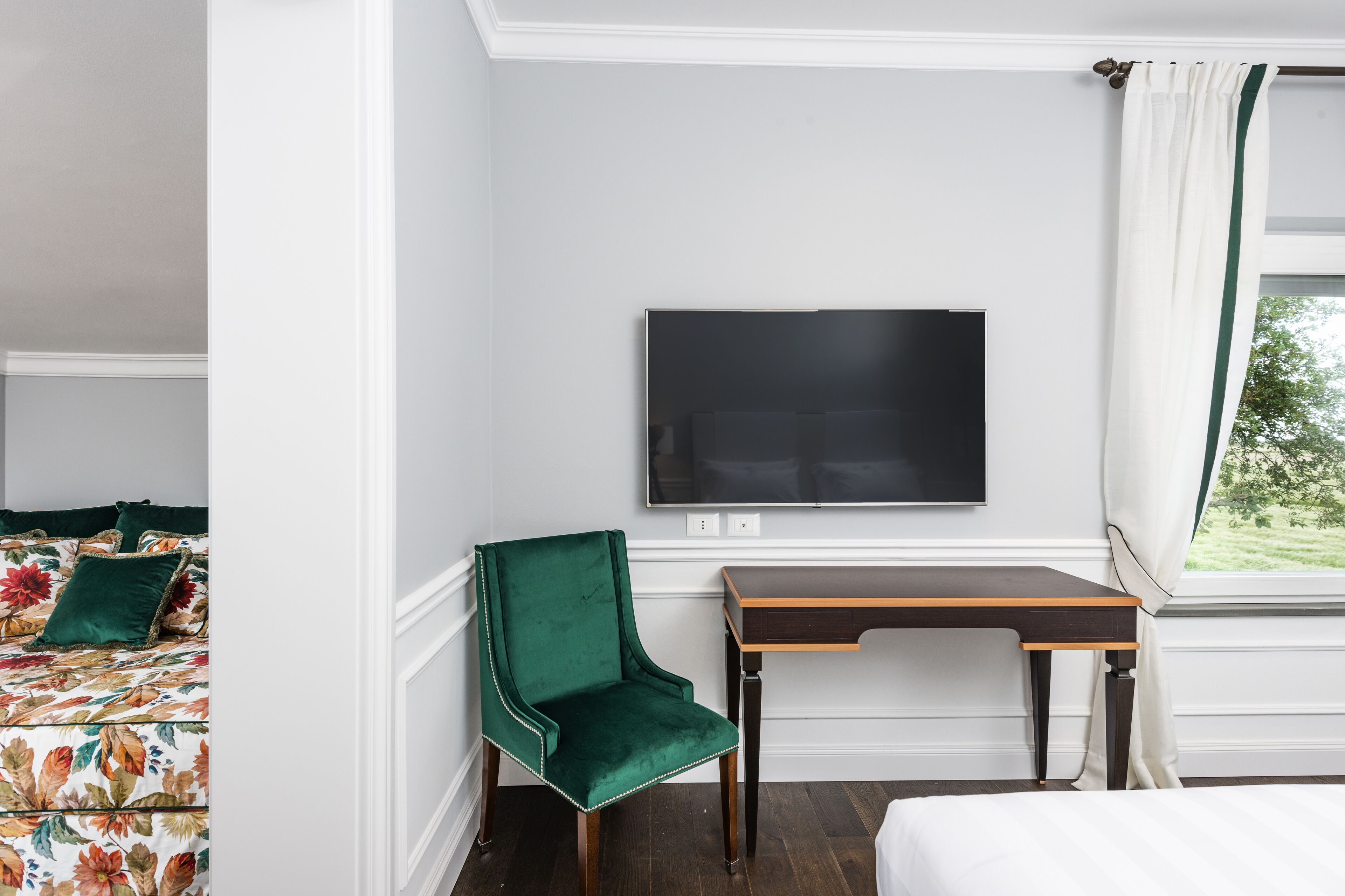 A hotel room with a bed, a green upholstered chair, a wooden desk, and a flat-screen TV mounted on the wall.