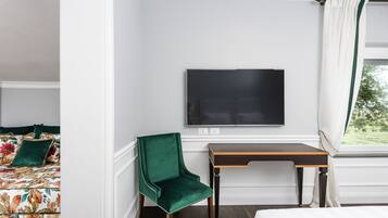 Une chambre d’hôtel avec un lit, un fauteuil recouvert d’un tissu vert, un bureau en bois et un téléviseur à écran plat fixé au mur.