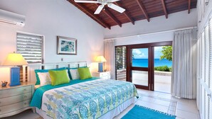 Een slaapkamer met een bed, nachtkastjes, een raam met jaloezieën, een plafondventilator en uitzicht op de oceaan.