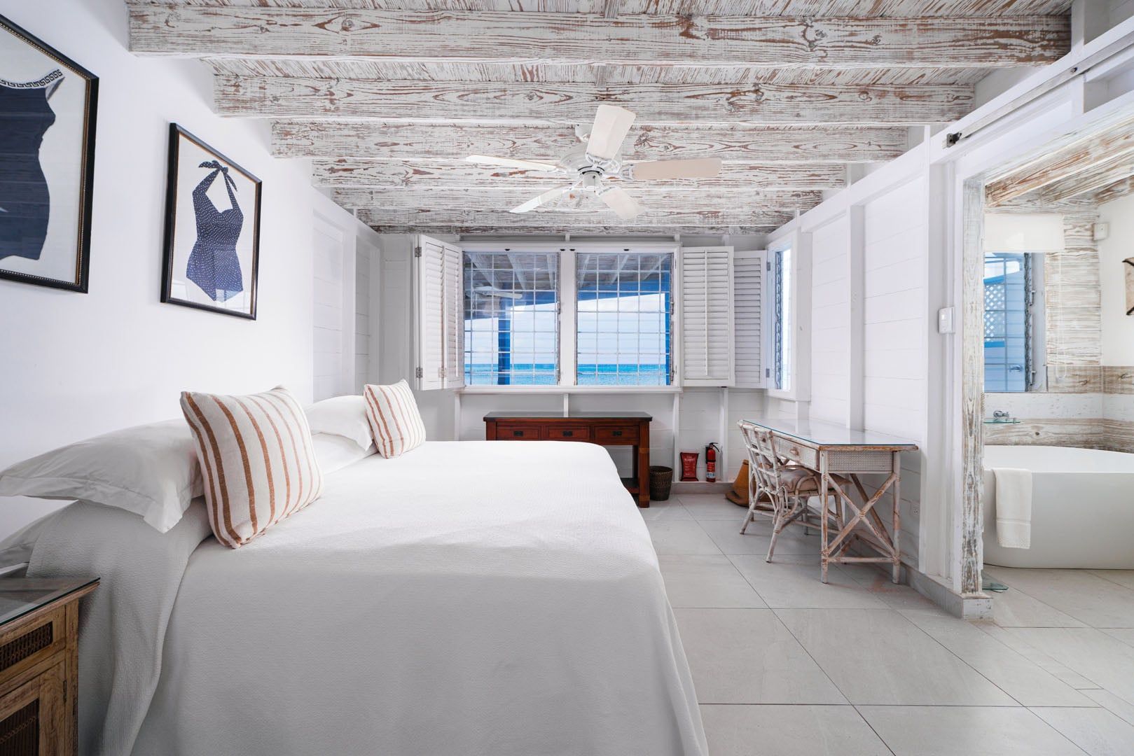 A bedroom with a large bed, a wooden ceiling, a window with shutters, and a bathroom with a bathtub.