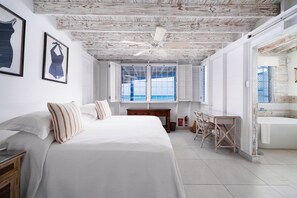 A bedroom with a large bed, a wooden ceiling, a window with shutters, and a bathroom with a bathtub.