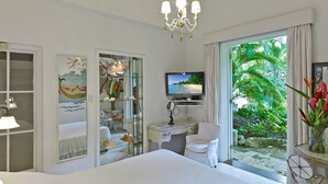 A hotel room with a bed, a desk, a TV, a chair, a bookshelf, and a large window with a view of greenery.