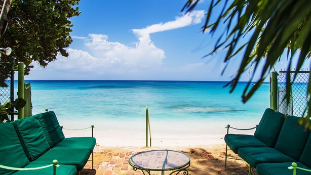 A beachside seating area with a table and two green sofas, overlooking a clear blue sea.