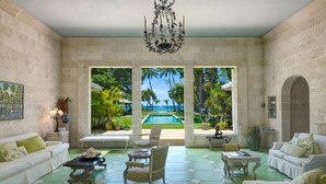 A spacious living room with a view of a pool and palm trees, furnished with white sofas and a chandelier.