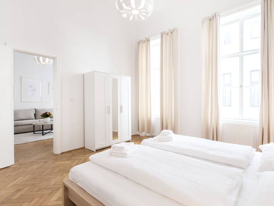 A modern hotel room with two beds, a wardrobe, a sofa, and a view of the city.