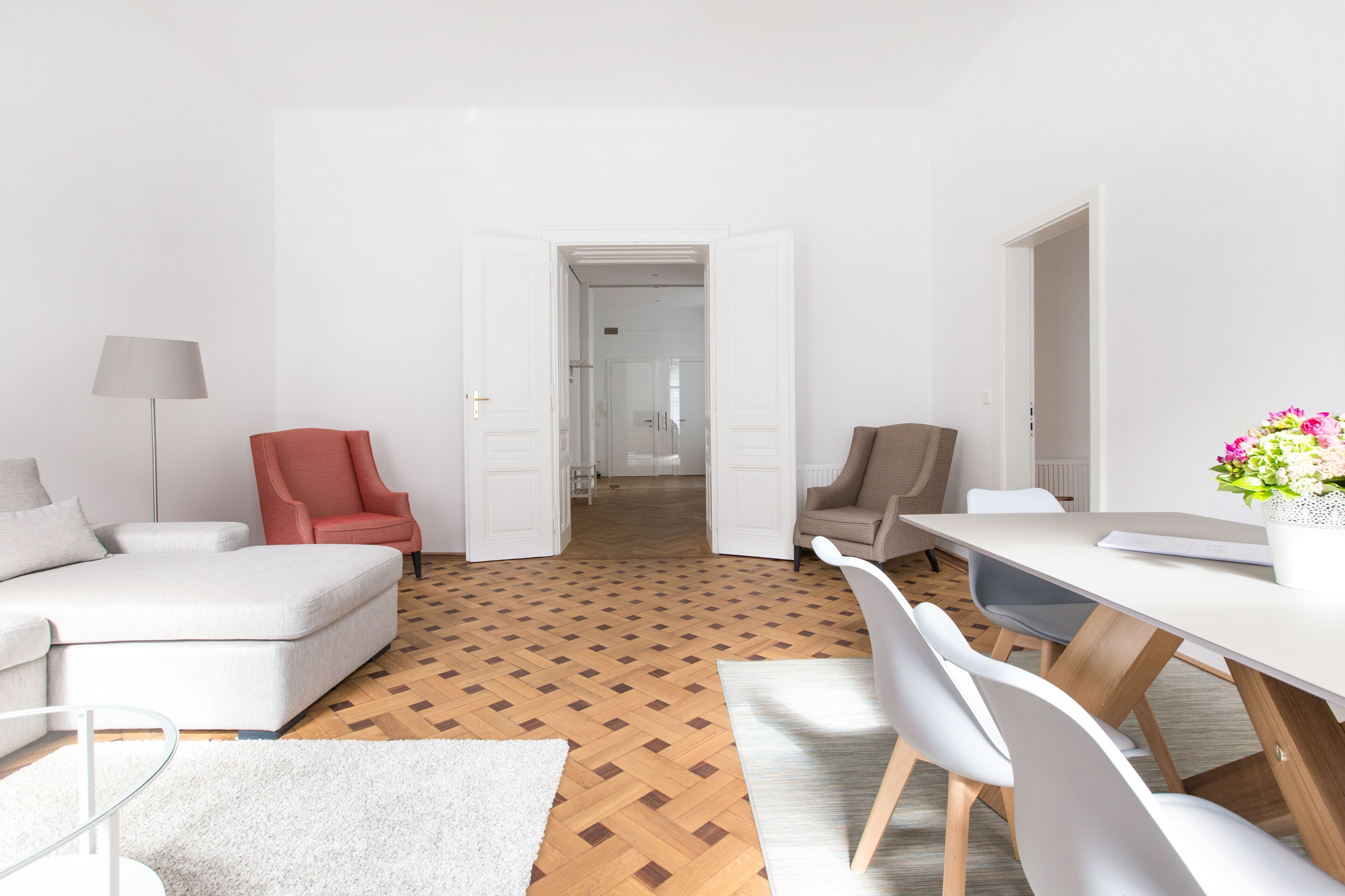 Ein modernes Wohnzimmer mit Holzboden, einem weißen Sofa, einem roten Sessel und einem Essbereich mit weißem Tisch und Stühlen.