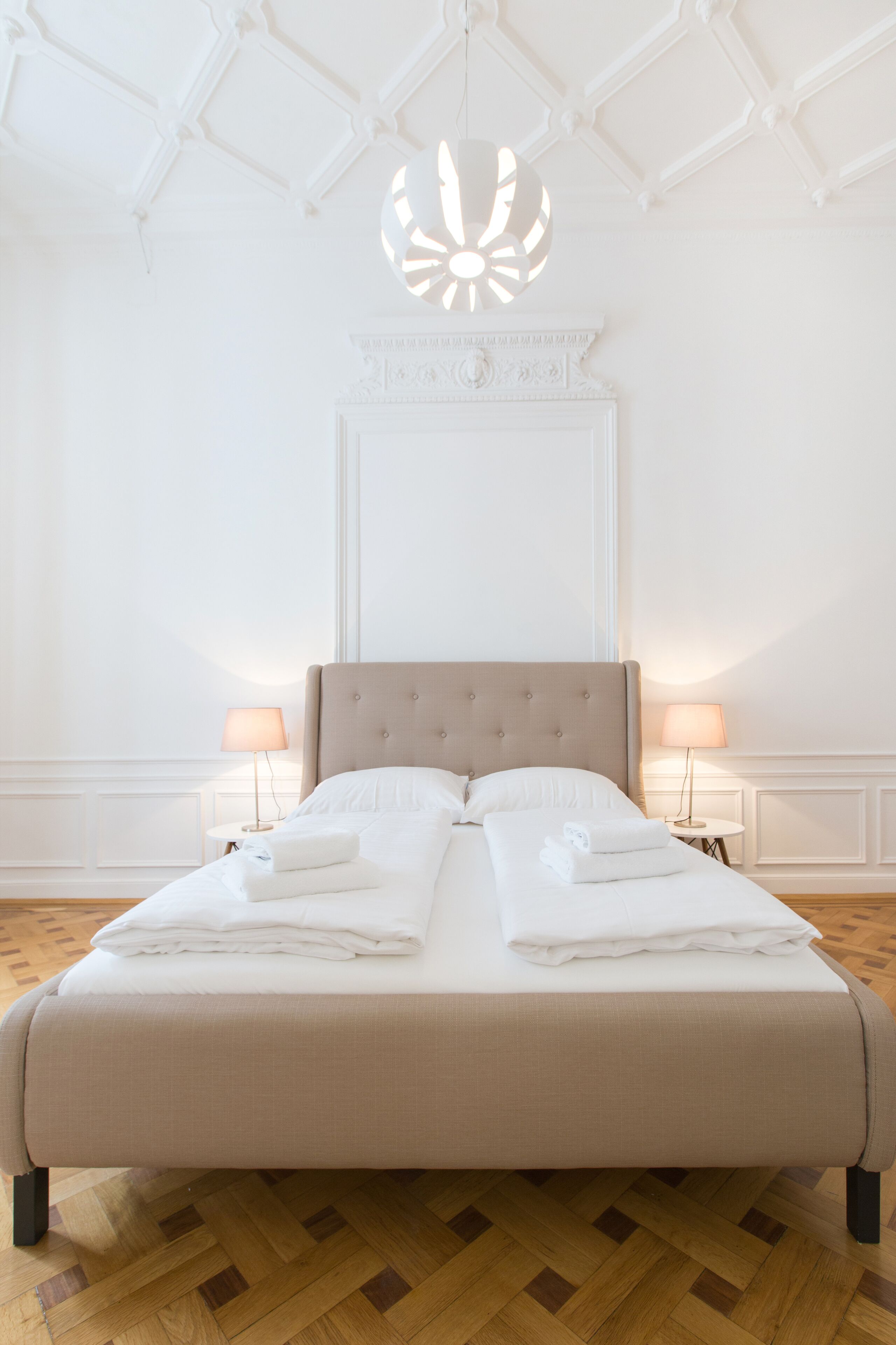 A neatly made bed with white linens and towels, a tufted headboard, and two bedside lamps on a wooden parquet floor.