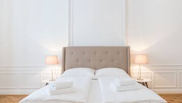 A neatly made bed with white linens and towels, a tufted headboard, and two bedside lamps on a wooden parquet floor.