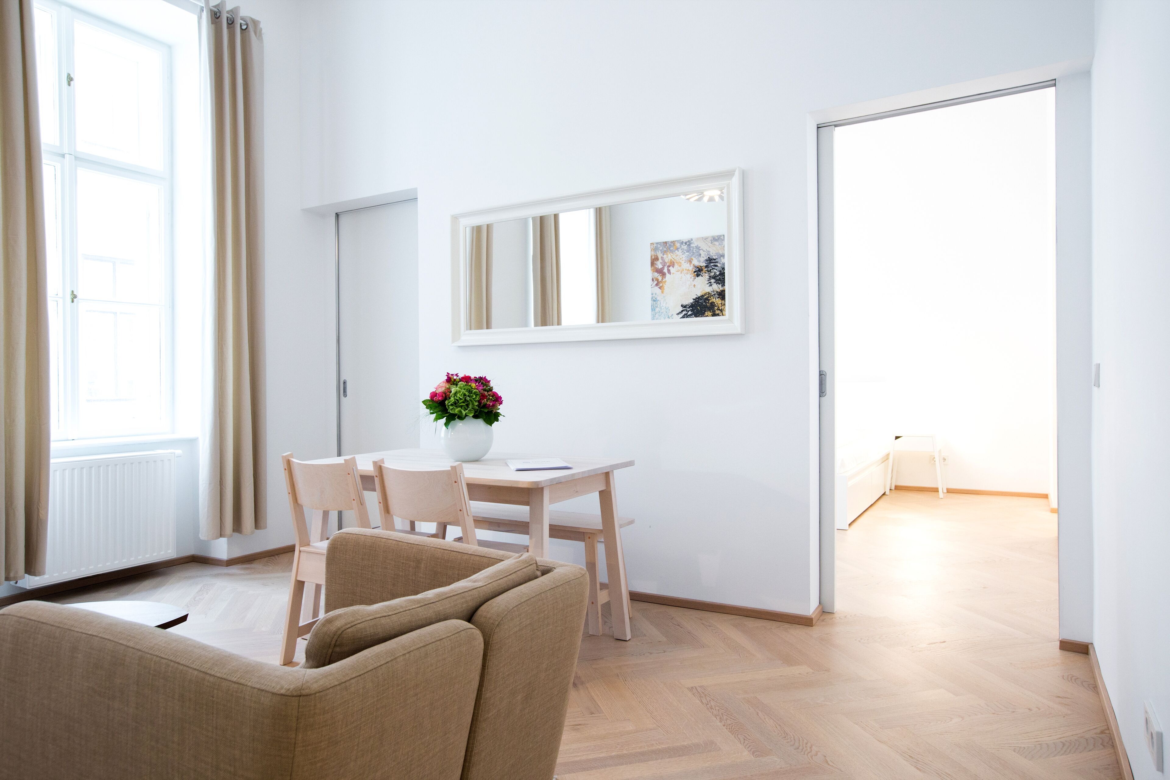 A modern living room with a sofa, a dining table with chairs, a vase of flowers, and a large mirror on the wall.