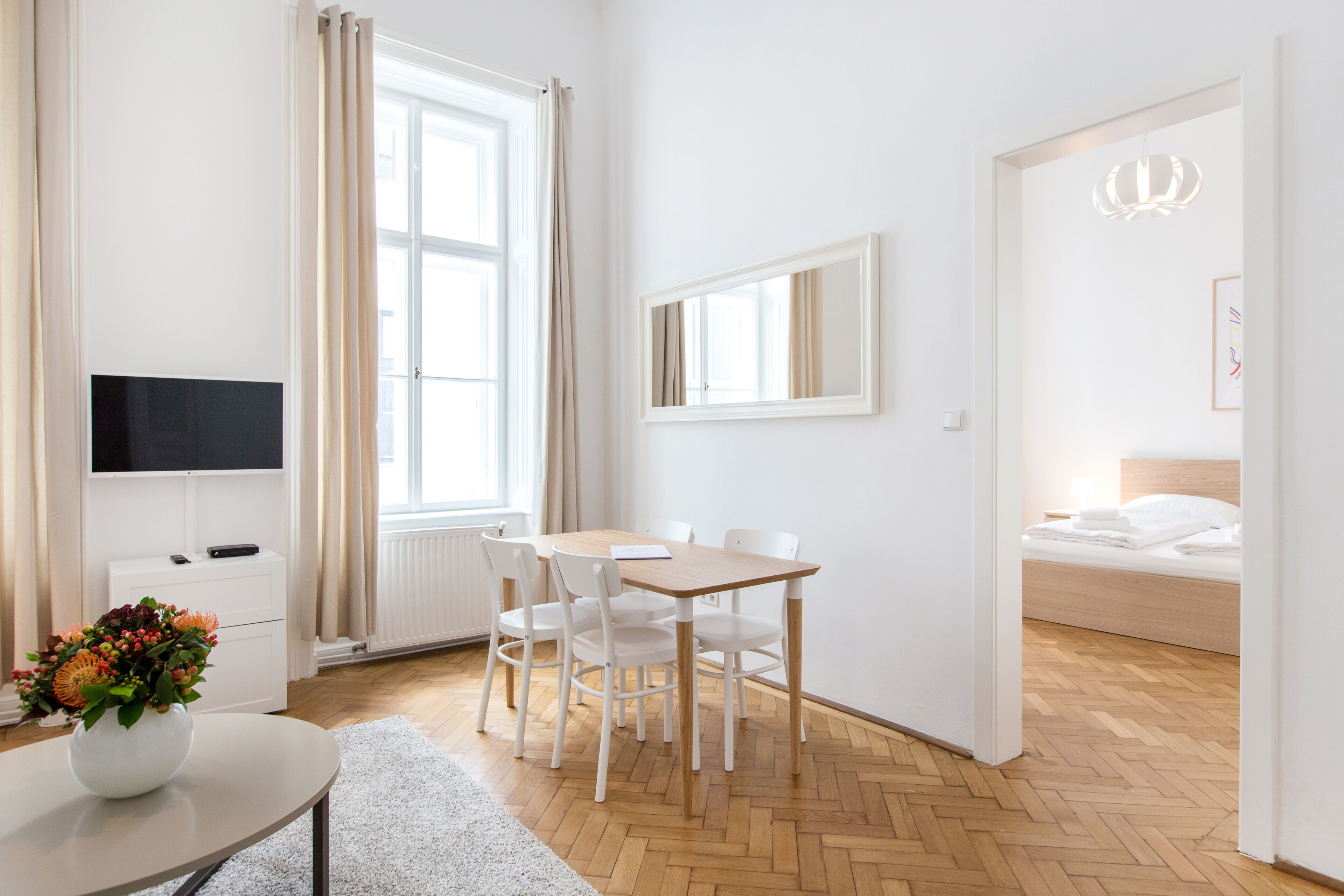 A modern hotel room with a wooden herringbone floor, a dining area with a table and chairs, a flat-screen TV, a bedside table with a vase of flowers, and a bed with white bedding.