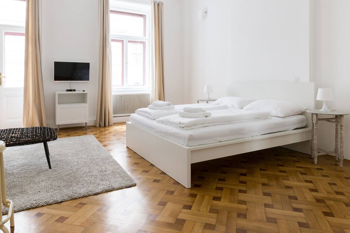Hotellihuone, jossa on puulattia, valkoinen sänky, valkoisella jalustalla oleva televisio ja pieni pöytä, jossa on lamppu.