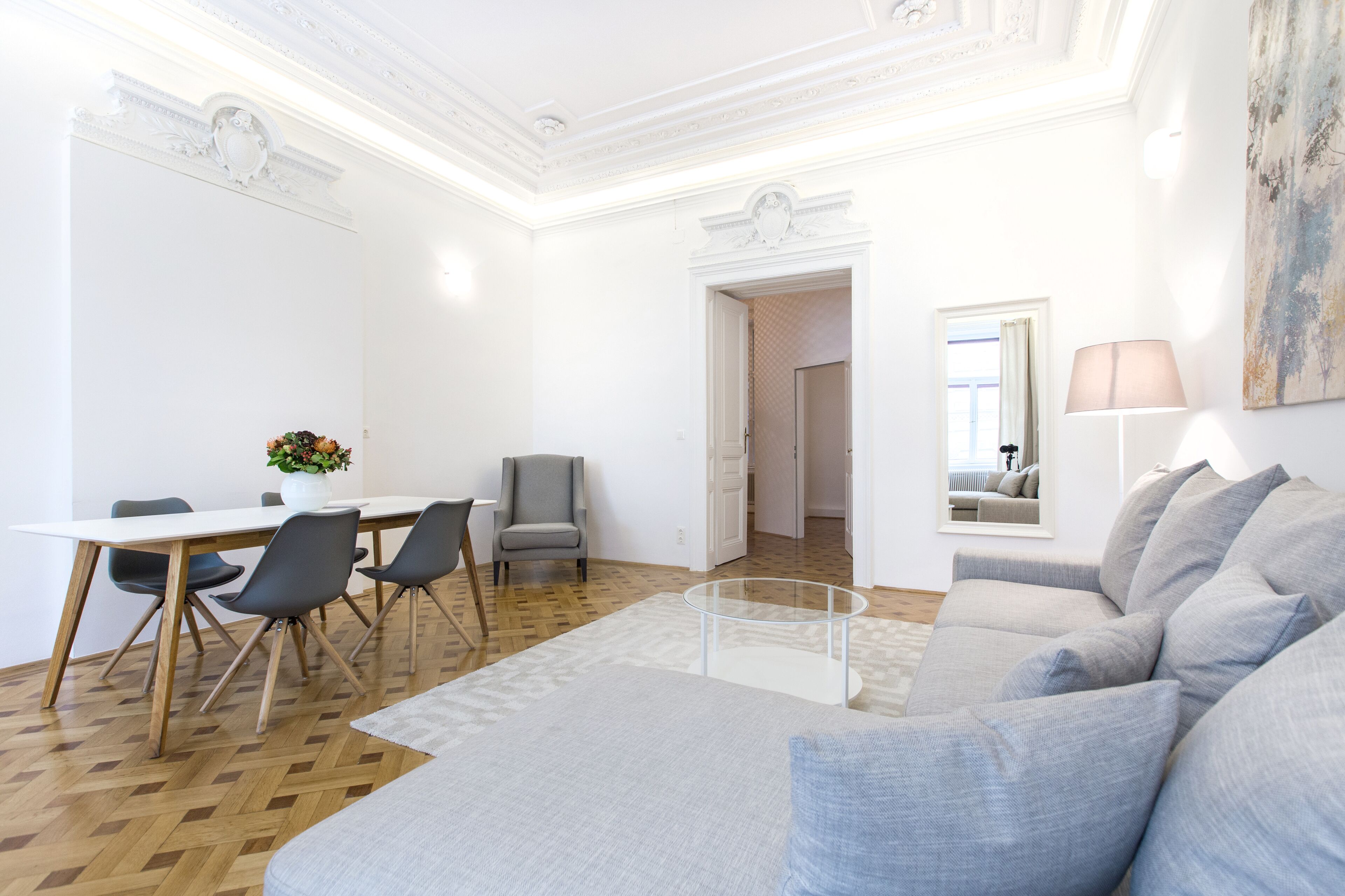 A modern living room with a grey sofa, a glass coffee table, and a dining area with a white table and chairs.