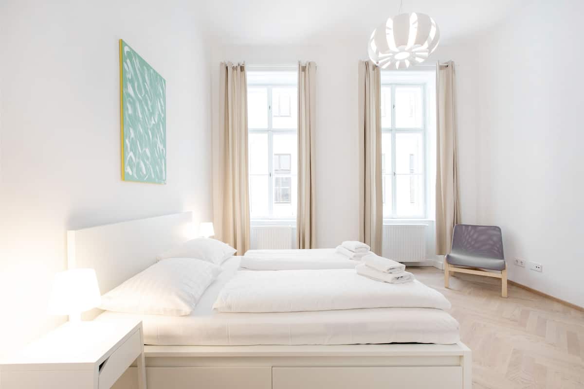 Moderni hotellihuone, jossa on suuri sänky, kaksi ikkunaa, tuoli ja riippuvalaisin.