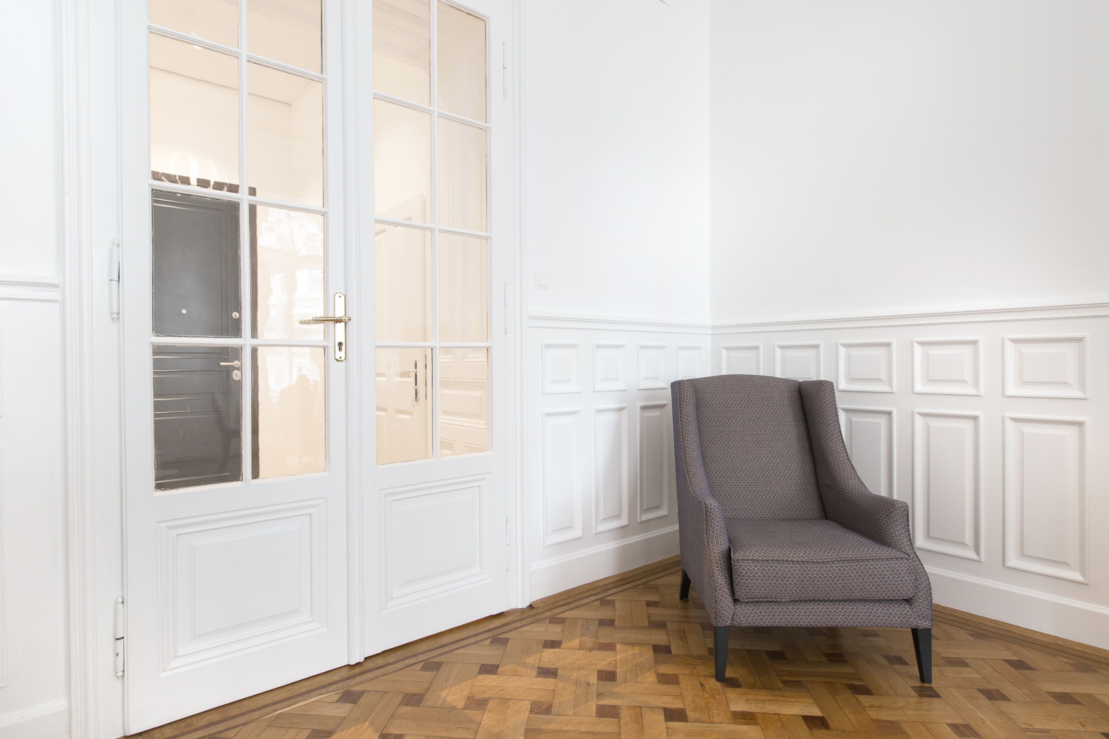 A room with a wooden parquet floor, a white door with glass panels, and a grey upholstered armchair.