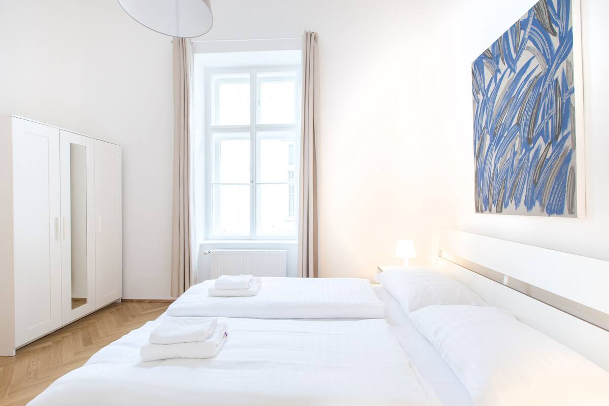 Hotellihuone, jossa on kaksi sänkyä, suuri ikkuna, vaatekaappi ja taulu seinällä.