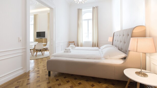 A hotel room with a large bed, a bedside table, a lamp, and a view of the dining area. - HeyMi Apartments in City Center (Vienna)