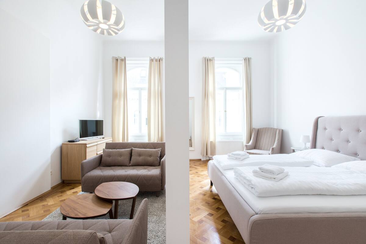 Moderni hotellihuone, jossa on sohva, pieni pyöreä pöytä, siististi taitelluilla pyyhkeillä varustettu sänky ja televisio, joka on sijoitettu puiseen kaappiin.