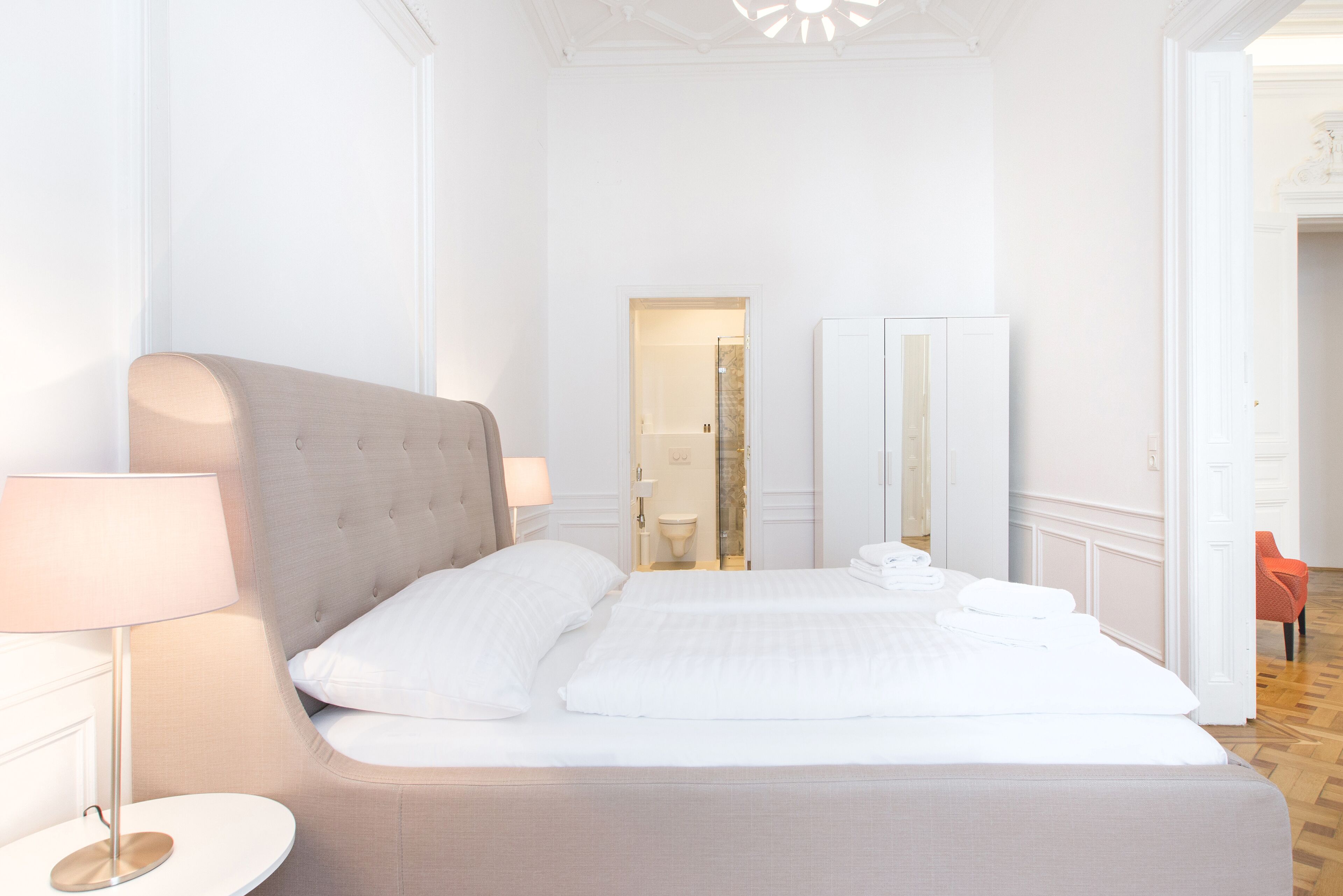 Ein Hotelzimmer mit einem großen Bett, einem runden Beistelltisch, einer Lampe, einem Spiegel und einer Tür zu einem anderen Raum.