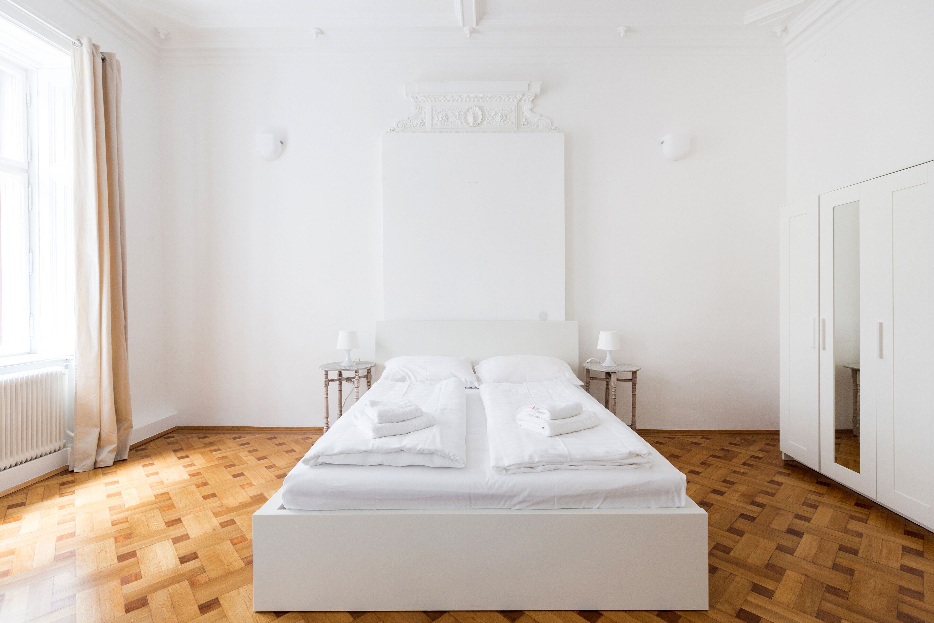Ein großes, ordentlich bezogenes Bett mit weißen Leinen und Kissen, zwei Nachttische mit Lampen, eine Heizung und ein Kleiderschrank mit Spiegel.